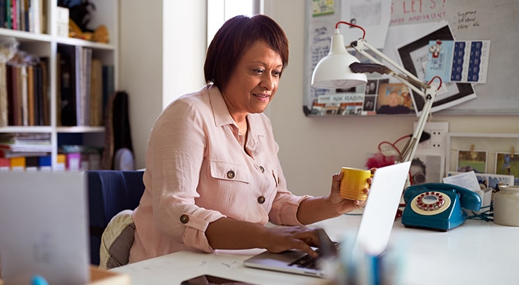 A woman in her home office, typing on the laptop and enjoying a coffee
